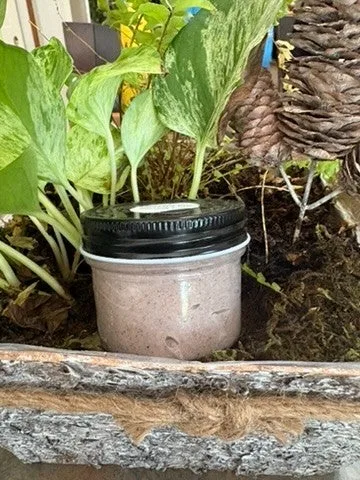 A jar of rose-tinted Inanna's body butter with a black lid among garden leaves and pinecones