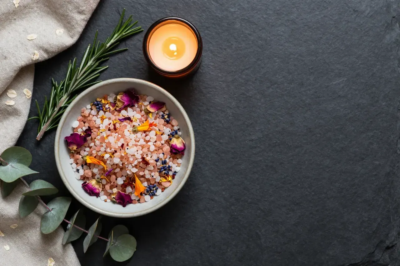 A bowl of pink Himalayan and white sea salts scattered with rose petals, lavender and calendula, beside a lit candle on dark slate